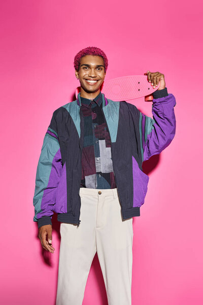 good looking man in bold stylish attire posing unnaturally with skateboard on pink backdrop