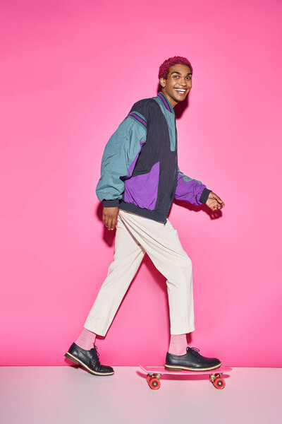cheerful young male model posing on skateboard in profile and smiling at camera on pink backdrop