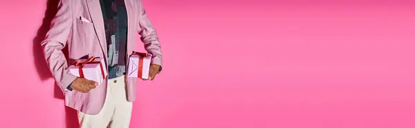 stock image cropped view of stylish young man in vibrant outfit with presents in hands on pink backdrop, banner