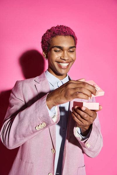 cheerful young man with silver accessories in pink blazer posing with small gift on pink backdrop