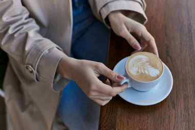 Trençkotlu genç bir kadının elinde kapuçino ve açık hava kahvesi, latte sanatı.