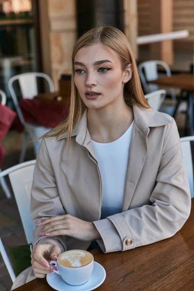 young blonde woman in trench coat enjoying her cup of cappuccino outdoors in cafe, latte art