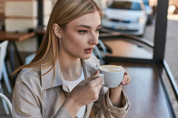 stock image dreamy young woman in trench coat enjoying her cup of cappuccino while sitting outdoors in cafe