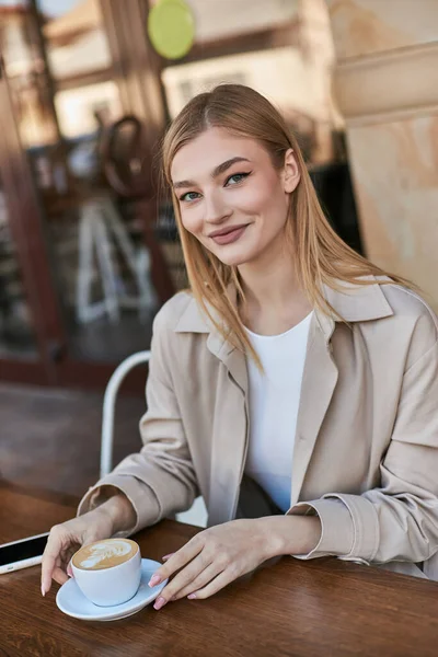 happy young woman in trench coat enjoying cup of cappuccino while sitting near smartphone in cafe