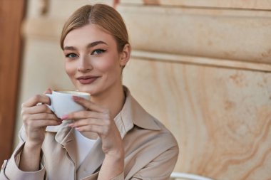 Süslü trençkotlu neşeli sarışın kadın kafede otururken elinde bir fincan kahve, içecek.