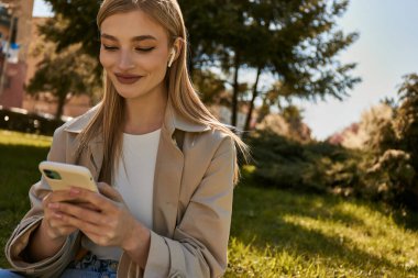 Kablosuz kulaklıklı ve şık trençkotlu mutlu sarışın kadın parkta akıllı telefonunu kullanıyor.