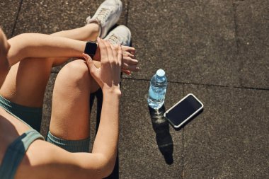 Aktif giyimli sporcu kadın, akıllı telefon ve su şişesinin yanındaki spor takip cihazının en üst görüntüsü.