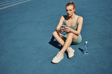 athletic woman in activewear sitting next to bottle of water and using mobile phone after workout clipart