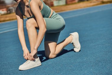 Koşu parkurunda koşmadan önce beyaz spor ayakkabılarıyla bağcıklarını bağlayan aktif giysili bir kadın.