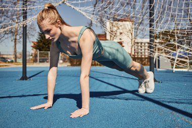 blonde young sportswoman with ponytail exercising in activewear near net outdoors, doing press ups clipart