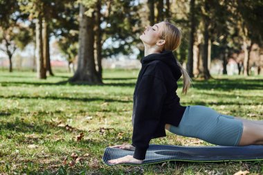 Sarı saçlı, spor giyinen, meditasyon yapan ve parkta yoga minderinde gerinen genç bir kadın.