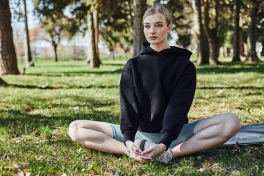 sportive young woman with blonde hair and sportswear sitting on mat and meditating in park clipart