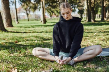 sarışın ve spor giyimli genç bir kadın spor minderinde oturuyor ve parkta meditasyon yapıyor.