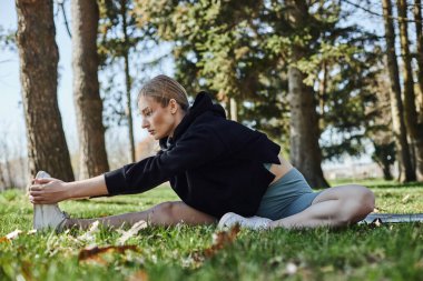 Parktaki spor kıyafetleri ve sarı saçlı formalı genç bir kadın spor kıyafetiyle spor minderinde otururken bacak esnetiyor.