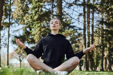 Sarı saçlı ve spor giyimli güzel bir kadın parkta meditasyon yaparken mindere oturuyor.