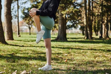 Spor kıyafetli genç bir kadının parkta çimlerin üzerinde durarak bacaklarını esnetirkenki görüntüsü.