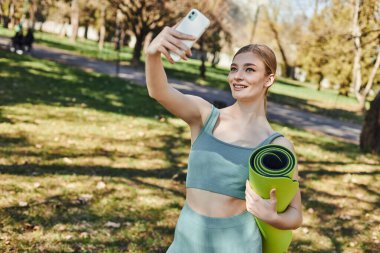 Aktif giyinen mutlu sporcu kadın akıllı telefondan selfie çekiyor ve parkta spor paspası tutuyor.