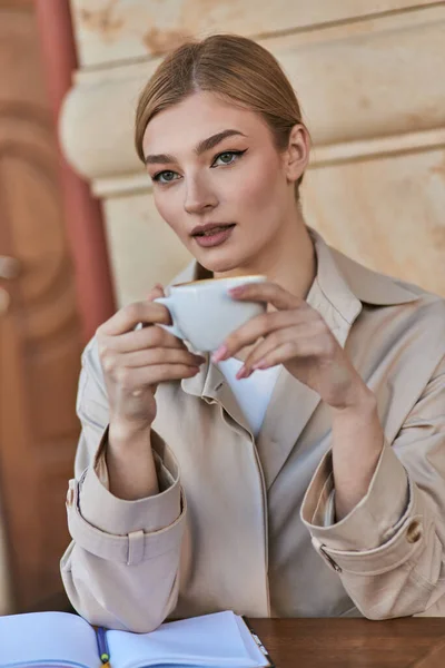 stock image elegant blonde woman in stylish trench coat holding cup of coffee while sitting in cafe, beverage