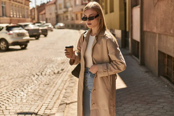blonde woman in trendy trench coat holding coffee to go while walking on street in sunny day