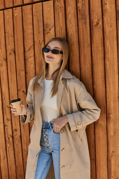 happy woman in trench coat and sunglasses holding coffee to go and standing with hand in pocket