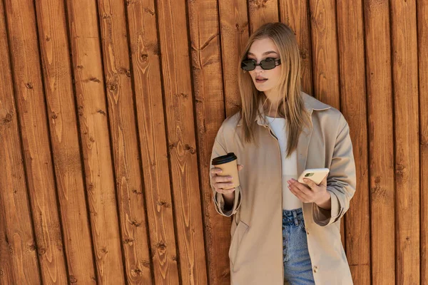 stylish woman in trench coat and sunglasses holding takeaway coffee and using smartphone near fence