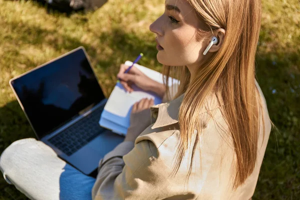 Молодая Блондинка Плаще Пишет Блокноте Используя Ноутбук Сидя Газоне Парке — стоковое фото