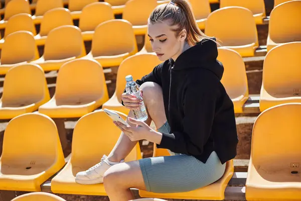 在体育场锻炼后 身穿马尾辫的女运动员拿着一瓶水 用智能手机 — 图库照片