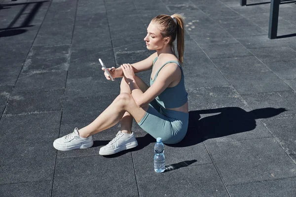 stock image fit woman in tight activewear sitting next to bottle of water and using smartphone after workout