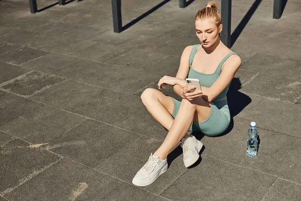 stock image sportswoman in tight activewear sitting next to bottle of water and using smartphone after workout