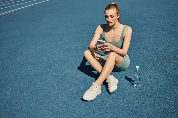 athletic woman in activewear sitting next to bottle of water and using mobile phone after workout