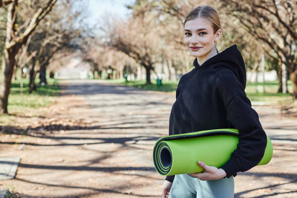 блондинка и стройная молодая женщина в черной спортивной толстовке держит фитнес коврик после тренировки в парке