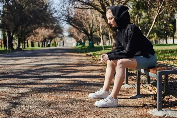 blonde and sporty woman in black sporty hoodie and cycling shorts sitting on wooden bench in park