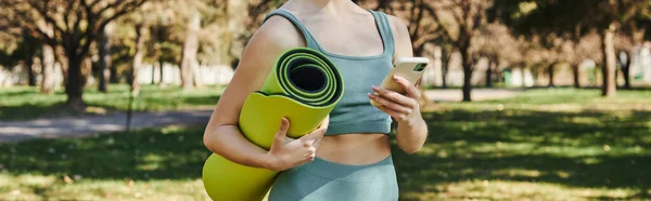 cropped banner of sportswoman in active wear holding smartphone and fitness mat in green park