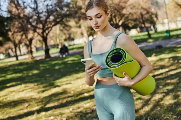 漂亮的女运动员穿着运动服 站在公园里用智能手机 拿着健身垫 — 图库照片