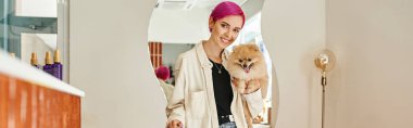 stylish woman with pomeranian spitz smiling at camera near reception desk in dog hotel, banner clipart