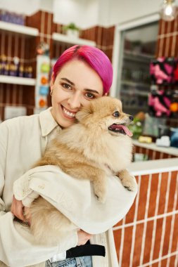 Mor saçlı mutlu kadın tüylü arkadaşını kucaklıyor ve köpek otelinde kameraya bakıyor.