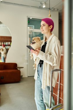 Küçük tüylü arkadaşı ve danışma masasının yanında akıllı telefonu olan ve köpek otelinde seyahat çantası taşıyan mutlu bir kadın.
