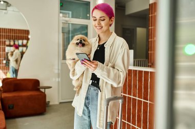 Küçük tüylü köpeği ve akıllı telefonu olan neşeli bir kadın resepsiyon masasının yanında ve köpek otelinde seyahat çantası taşıyor.