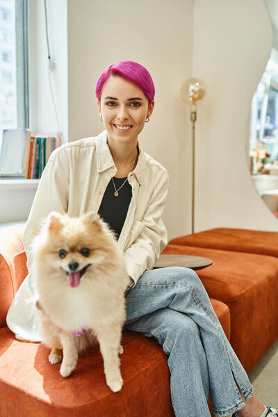 joyful purple-haired woman with pomeranian spitz sitting in reception area of modern pet hotel