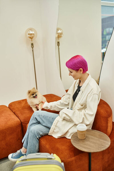 stylish woman playing with  own dog while sitting in pet hotel near travel bag and coffee to go