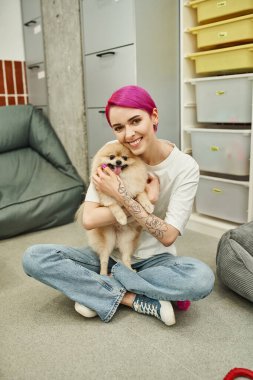 Dövmeli hayvan bakıcısı, mor saçlı, tüylü köpeğe sarılan ve evcil hayvan otelinde kameraya gülümseyen.
