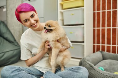 Neşeli ve şık köpek bakıcısı evcil oteldeki kameraya bakarken Pomeranian Spitz 'i kucaklıyor.