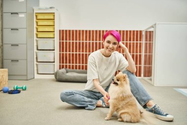 Sevgi dolu köpek bakıcısı komik Pomeranya tükürüğünün yanında oturmuş evcil hayvan otelinde kameraya gülümsüyor.