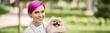 smiley and trendy woman with cute pomeranian spitz in hands looking at camera in park, banner clipart