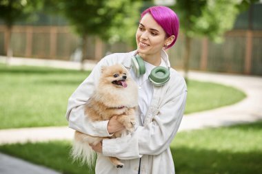 Gülümseyen, mor saçlı, kulaklıklı bir kadın. Elinde sevimli, saf bir köpekle dışarıda yürüyor.