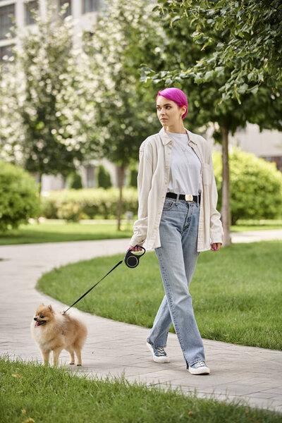 stylish woman looking away while walking in green city with pomeranian spitz on roulette leash