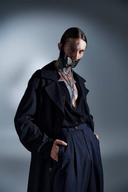 Siyah elbiseli, elleri ceplerinde dövmeleri olan çekici bir adamın dikey görüntüsü.