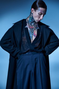 Siyah giyinmiş, elleri kalçasında, modaya uygun şekilde poz veren dövmeli genç bir adamın dikey çekimi.