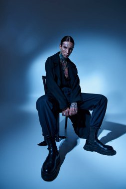 Sandalyede oturan dövmeli ve aksesuarlı yakışıklı genç adamın dikey çekimi, moda konsepti