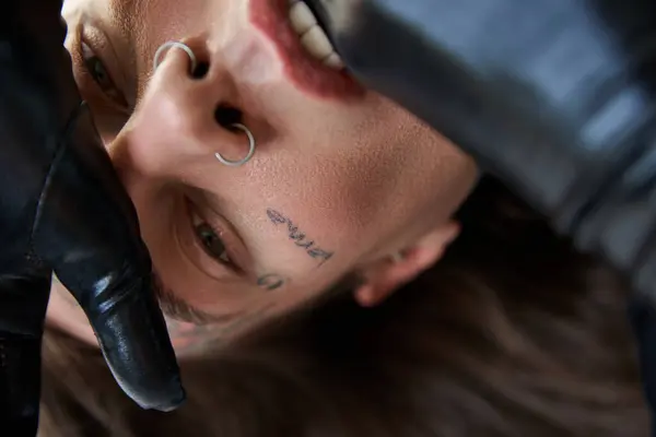 close up of handsome young man with tattoos on face and piercing in black gloves, fashion concept